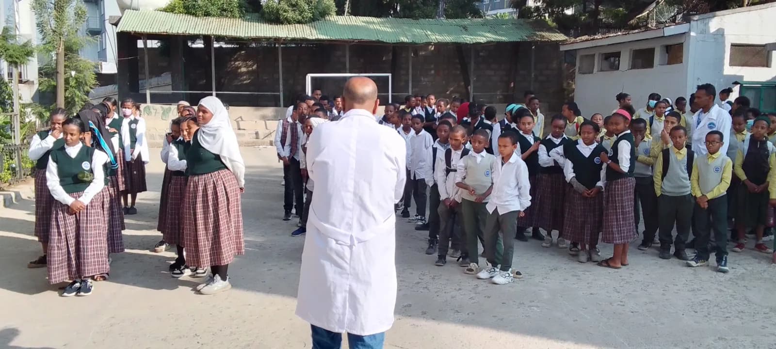 A physician in a white coat speaking to rows of students in a school courtyard